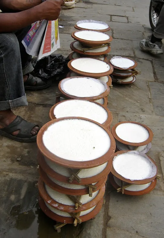 A street vendor selling Juju Dhau in a corner of busy Ason Tole chowk in Kathmandu. The yogurt containers (maato ko kataaro) are stacked high with the help of a triangular wooden frame stand.